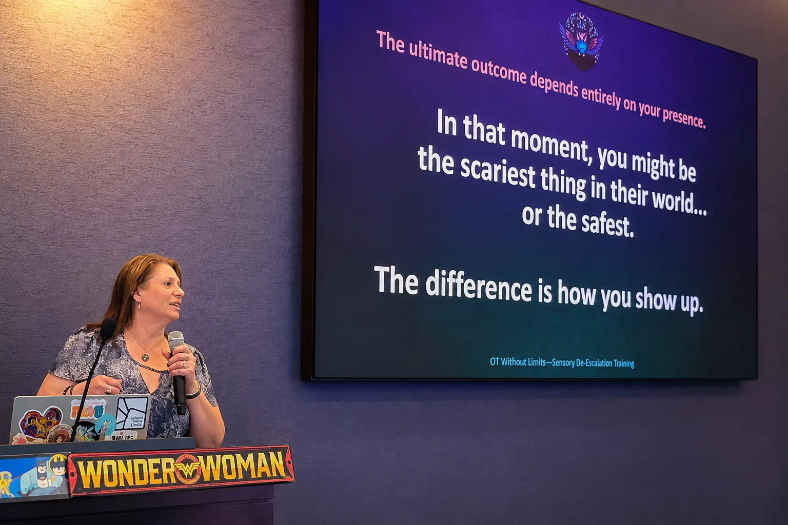 Dr Carrie Punger speaking into a microphone at a podium beside a large presentation screen displaying a quote about presence and sensory de-escalation training from OT Without Limits.