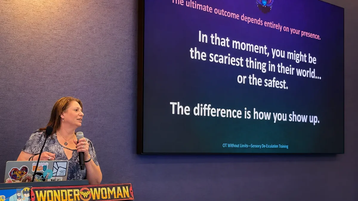 Dr Carrie Punger speaking into a microphone at a podium beside a large presentation screen displaying a quote about presence and sensory de-escalation training from OT Without Limits.